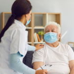 An elderly man is receiving medical attention from a nurse who is wearing personal protective equipment, including a mask.