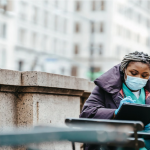 An image of a woman donning a face mask and face shield, sitting on a bench.