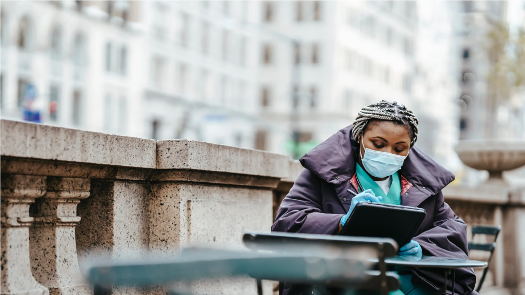 An image of a woman donning a face mask and face shield, sitting on a bench.